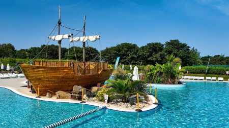 A wooden pirate ship model is anchored in a large outdoor pool surrounded by lush greenery under a clear blue sky in Cornwall.