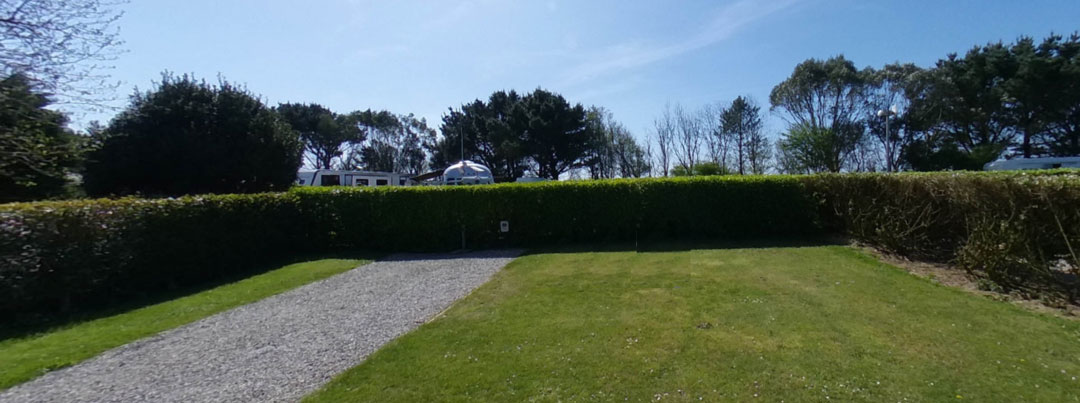 A sunny day in a Cornish holiday pitch with a grassy lawn and a gravel path. Tall, dense hedges surround the area, with trees and a blue sky in the background. In this idyllic holiday park setting, some structures, possibly tents or caravans, are visible beyond the hedge.