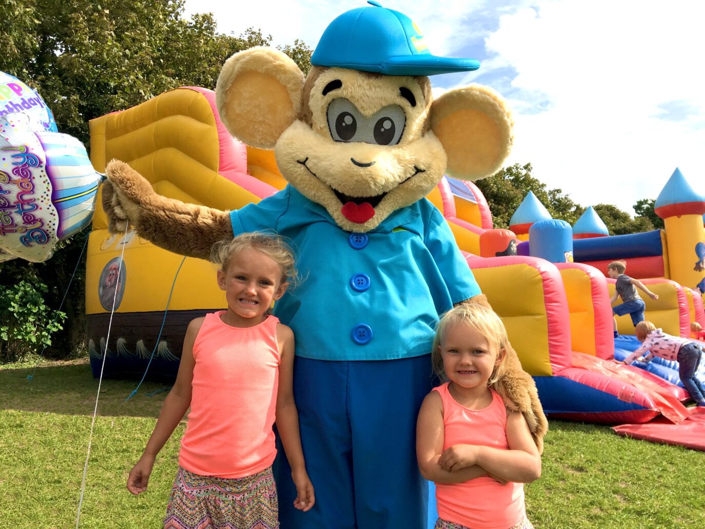 A person in a cartoonish mouse costume stands with two young girls at an outdoor birthday party at Monkey Tree Holiday Park in Cornwall, with a colorful inflatable bounce house in the background. The mouse is holding