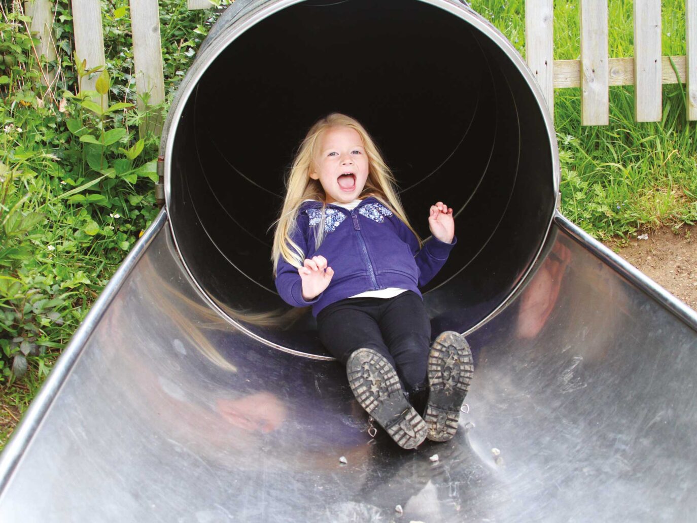 A young child with long blonde hair, wearing a purple hoodie and black pants, slides down a shiny metal tunnel slide, smiling with excitement. There is green grass and a wooden fence in the background.