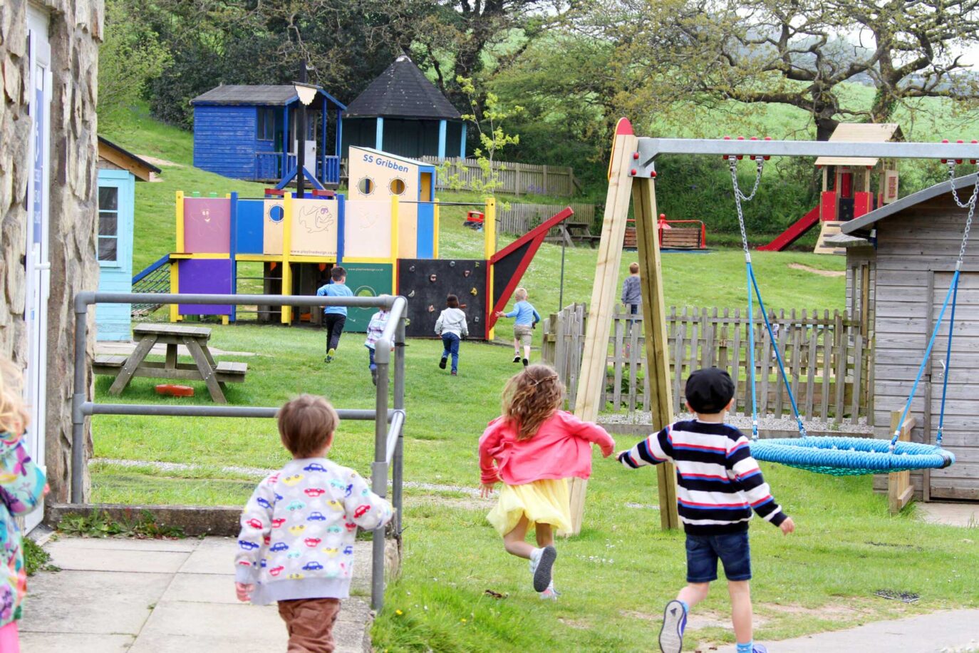 Children are playing in a vibrant playground near Newquay, where some kids dash towards a colorful play structure, while others enjoy the swings and wooden sheds. The scene captures the energy and joy of kids at Monkey Tree in Cornwall on a sunny day.