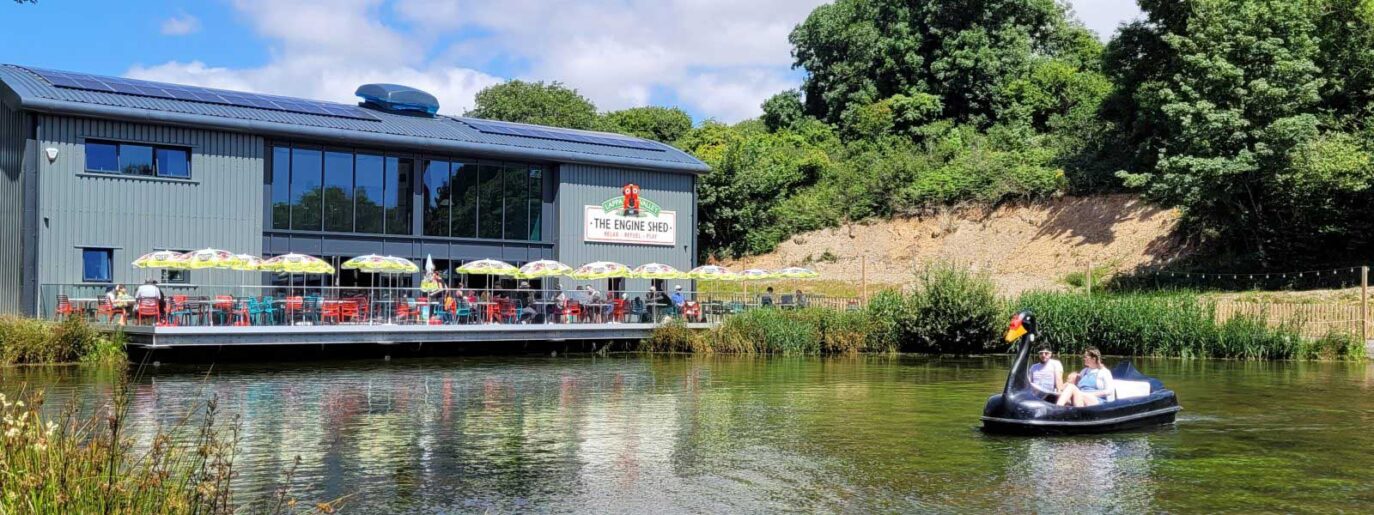 A large building with outdoor seating next to a pond. Two people are in a small boat on the water. Trees and a sunny sky are in the background.