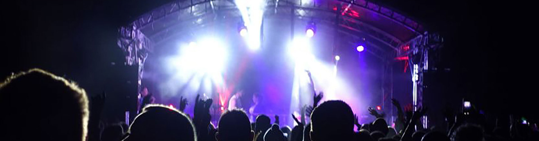 A lively outdoor concert scene at night in a holiday park, with a brightly lit stage and colorful spotlights. The silhouettes of a crowd with raised hands are visible in the foreground, creating an energetic and immersive atmosphere reminiscent of Cornwall's vibrant spirit.