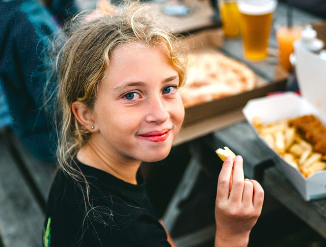 Young girl enjoying chips at an outdoor table with takeaway boxes of fish and chips and pizza in the background.