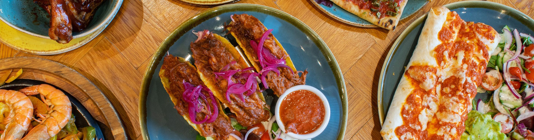 An assortment of colourful dishes including tacos with pickled red onions, a cheesy burrito with a side salad, prawns served with vegetables, and a selection of sauces on a wooden table.