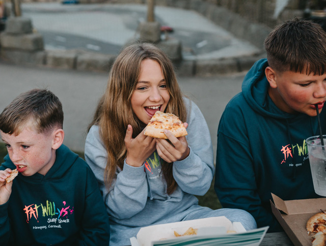 Three children enjoying pizza and drinks outdoors at Monkey Tree Holiday Park, with takeaway boxes on the table.