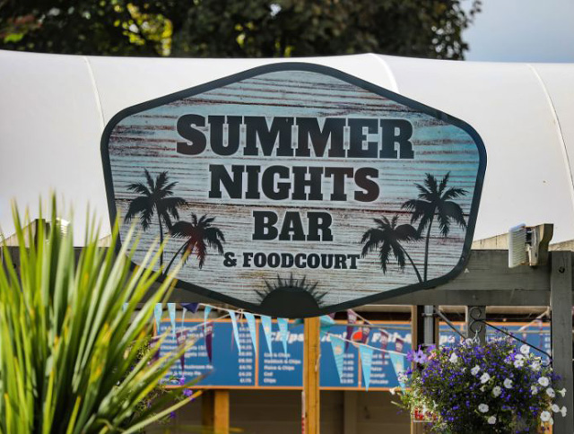 Sign for the Summer Nights Bar & Food Court at Monkey Tree Holiday Park, featuring a rustic wood background with palm tree silhouettes.