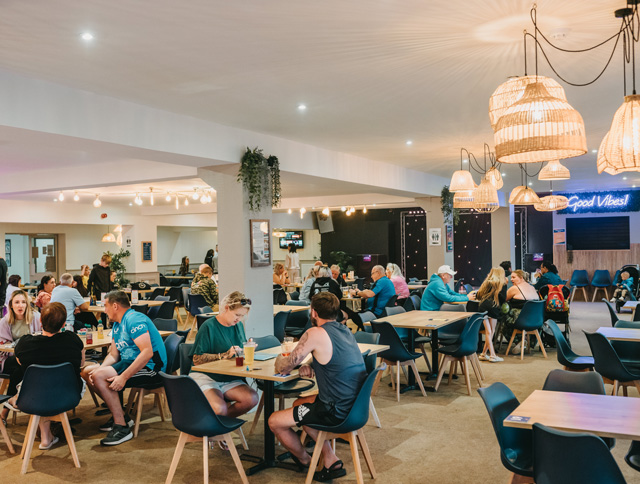 Guests dining and socialising inside The Beach Club at Monkey Tree Holiday Park, with modern decor, wooden tables, and ambient lighting.