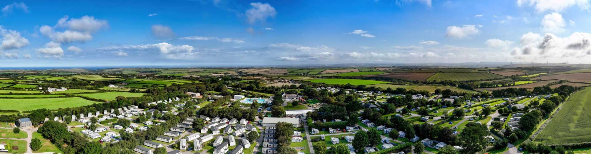 A panoramic view of a rural landscape featuring fields, trees, and a cluster of holiday homes and camping at the Monkey Tree Cornish holiday park. The sky is bright with scattered clouds, surrounded by lush greenery and farmlands stretching into the distance.