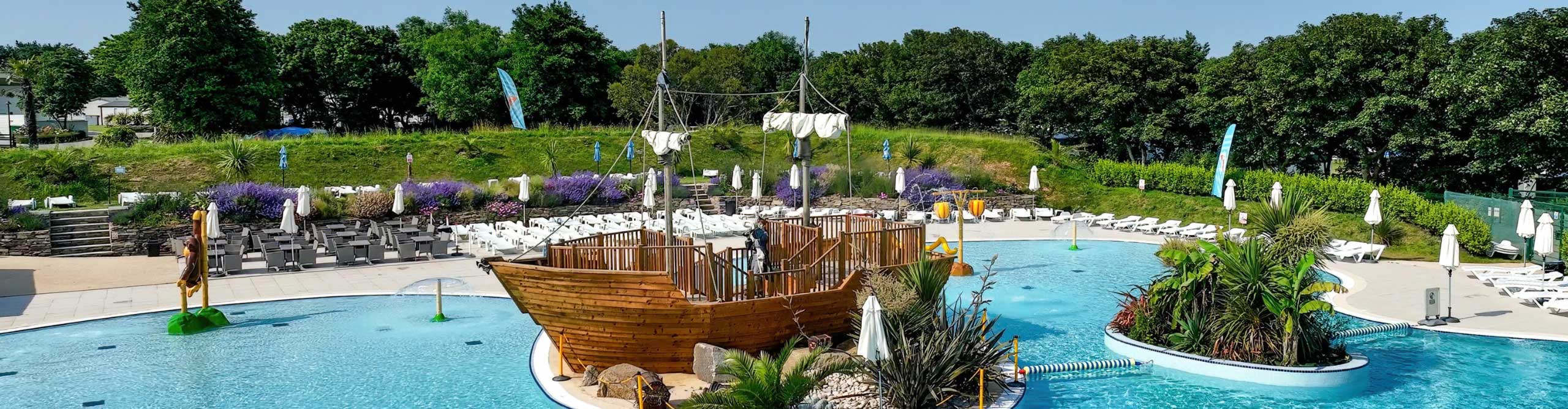 A large outdoor pool features a wooden pirate ship structure surrounded by lush greenery and sun loungers. The pool is decorated with tropical plants and has a clear blue sky overhead.